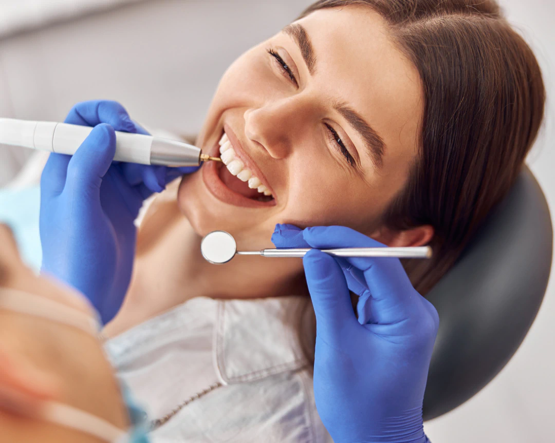 A smiling patient receives dental care from a dentist using a handheld dental drill and a mouth mirror.