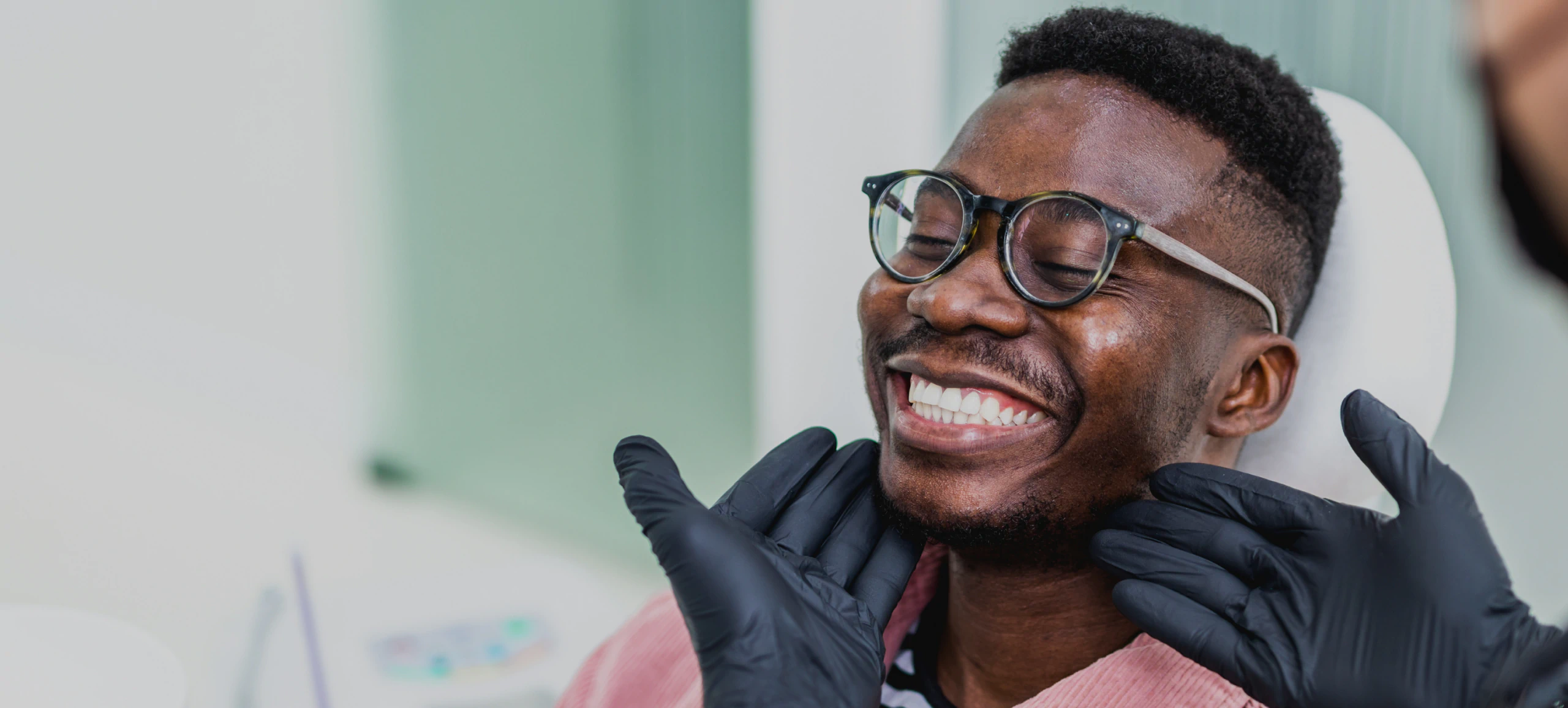 A smiling man in glasses has his jawline examined by a dental professional wearing black gloves.