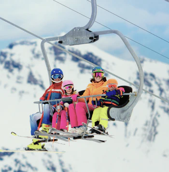 A family of four wearing ski gear and goggles rides a chairlift together with snowy mountains in the background.