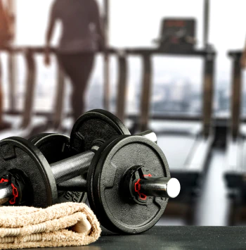 A pair of black dumbbells and a folded tan towel sit on a surface in a gym.